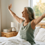 Woman sitting up in bed stretching in soft morning light, looking rested and energised.