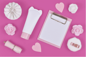 Flat lay with blank clipboard and various cute items like rose flowers, cream tube in shape of cat, paper hearts and powder puff on pink background