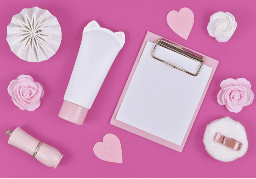 Flat lay with blank clipboard and various cute items like rose flowers, cream tube in shape of cat, paper hearts and powder puff on pink background